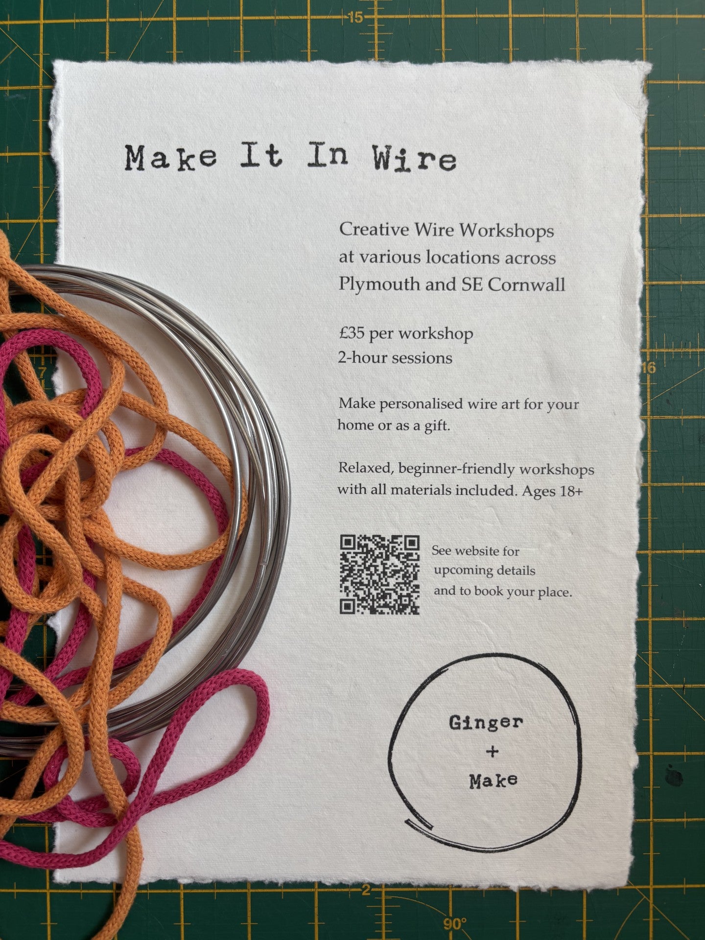 A printed workshop flyer titled 'Make It In Wire' laid on a green cutting mat, with coiled orange, pink, and silver wire draped across the left side. The flyer includes workshop details, a QR code, and the brand name 'Ginger + Make'.