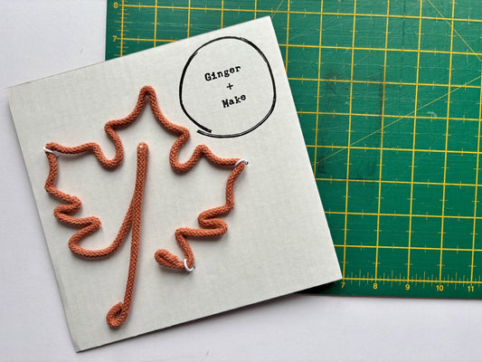 Knitted leaf patterns on a card with 'Ginger & Nake' branding, on a green cutting mat.