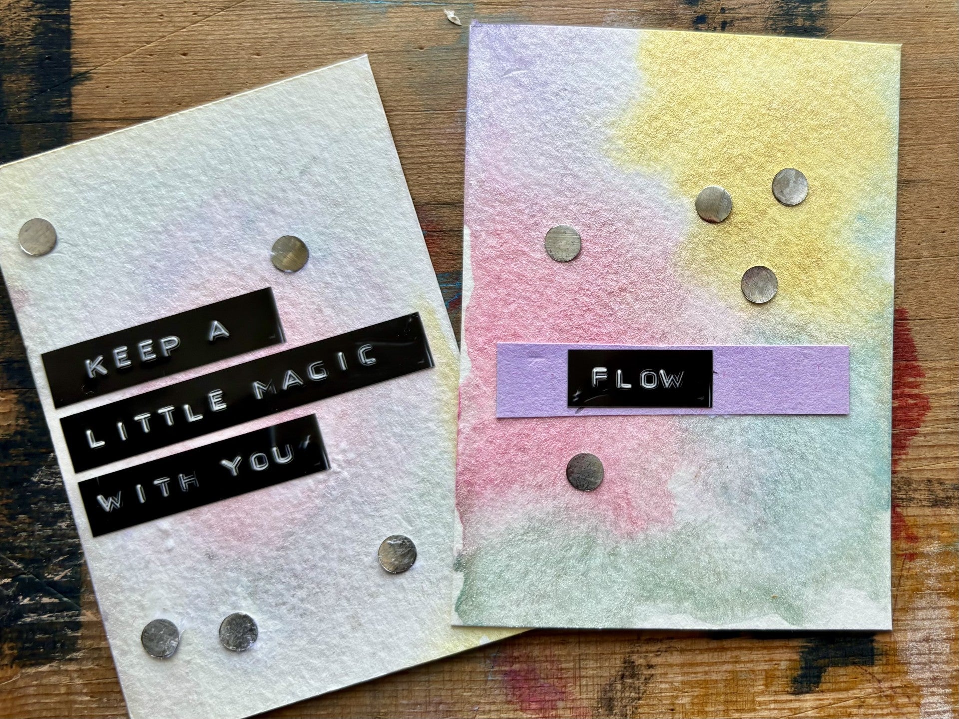 Two handmade cards with watercolor washes and small metallic coins, displayed on a wooden surface. One card is white with black text reading 'KEEP A LITTLE MAGIC WITH YOU', and the other is pastel pink, yellow, and blue with a purple banner reading 'FLOW'.