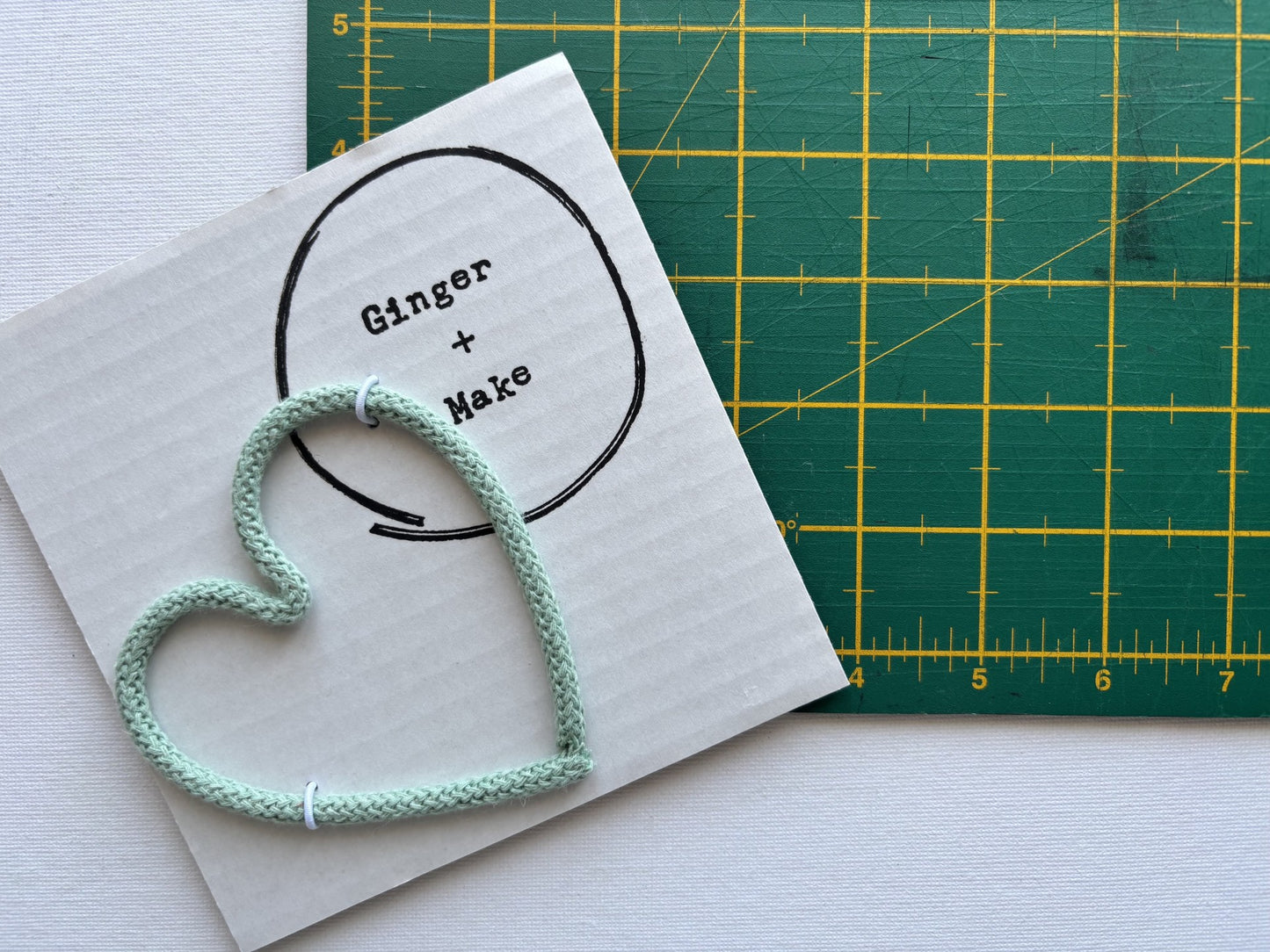 An aloe green knitted heart lies on white card printed with the words 'Ginger + Make' and a black circle outline. A green cutting mat with yellow grid lines is partially visible behind it on a white surface.