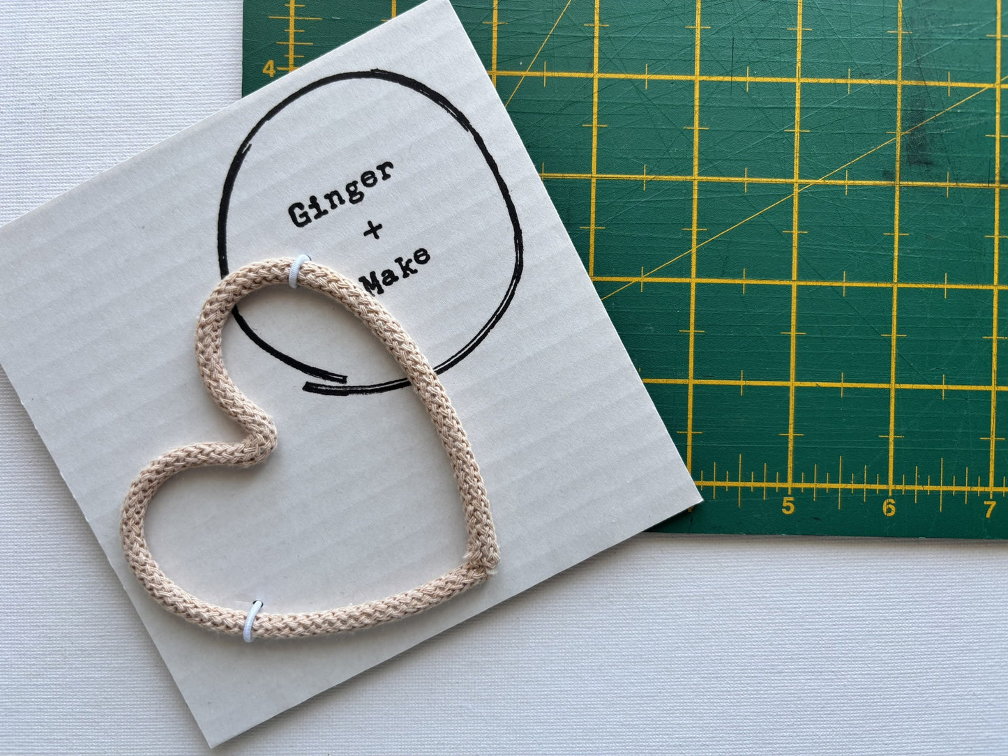 A handmade wire heart with biscuit cord, displayed on white card printed with 'Ginger + Make' next to a green cutting mat.