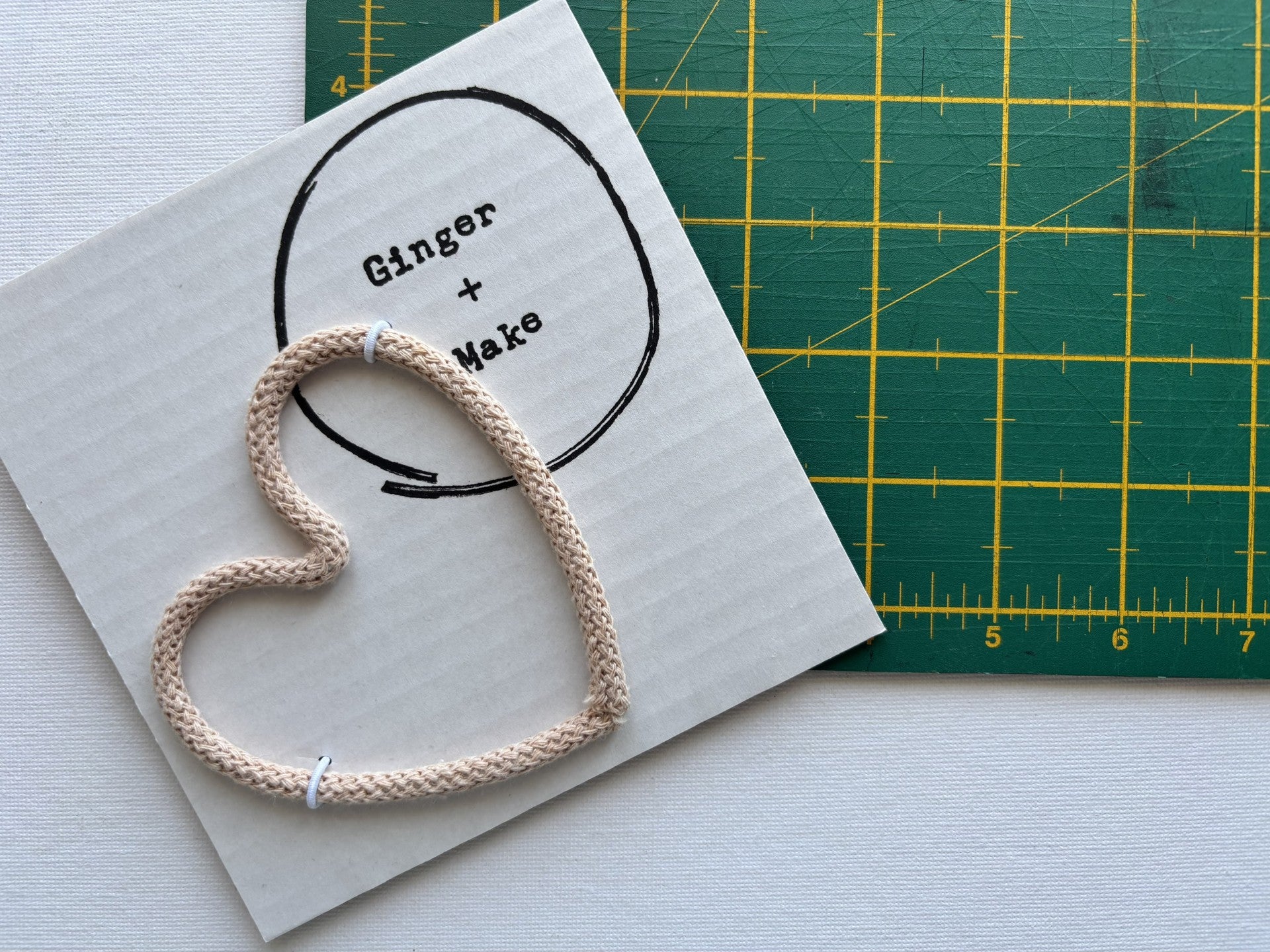 A handmade wire heart with biscuit cord, displayed on white card printed with 'Ginger + Make' next to a green cutting mat.