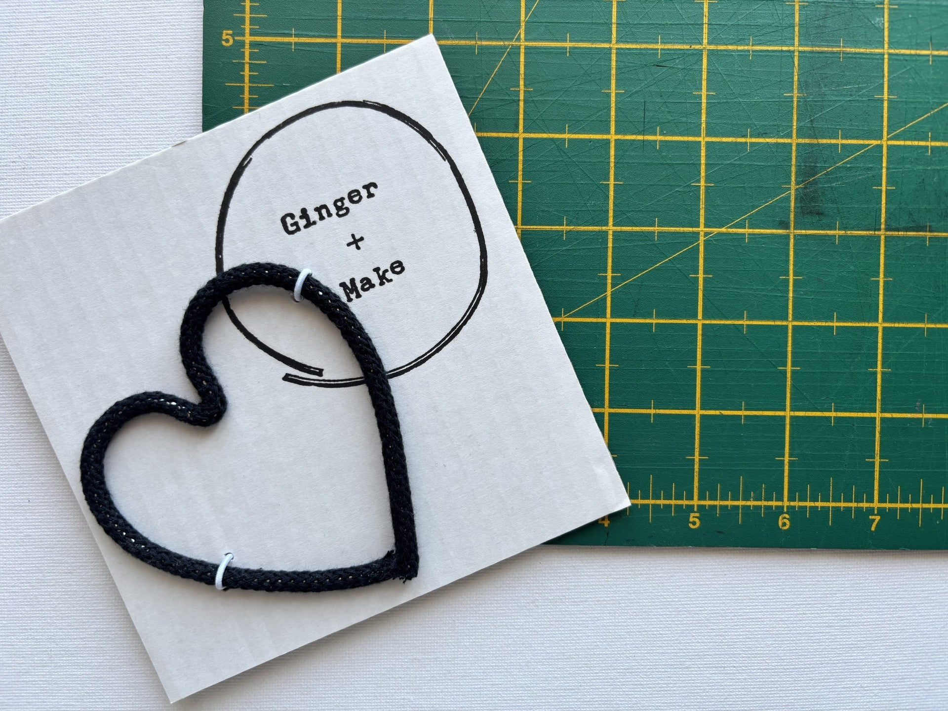 A handmade wire heart with black cord, displayed on white card printed with 'Ginger + Make' next to a green cutting mat.