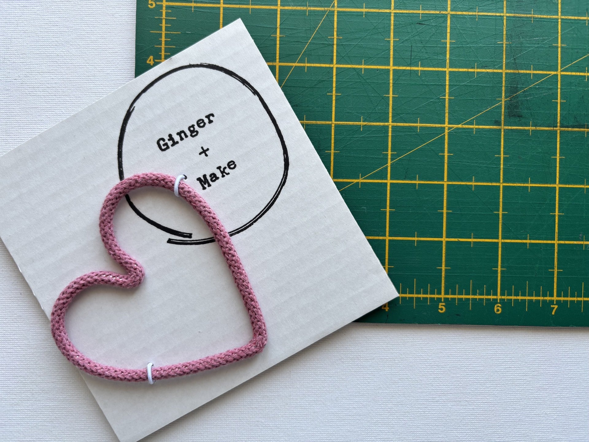 A handmade wire heart with pink cord, displayed on white card printed with 'Ginger + Make' next to a green cutting mat.