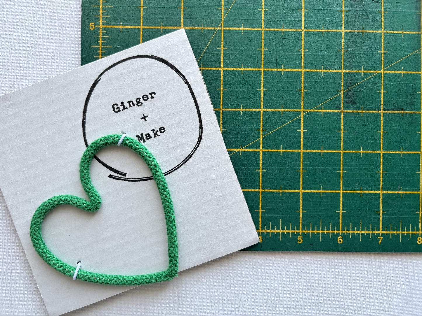 A handmade wire heart with green cord, displayed on white card printed with 'Ginger + Make' next to a green cutting mat.