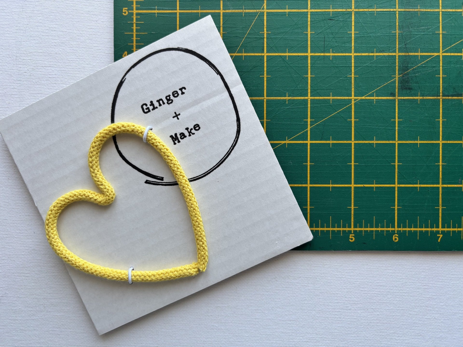 A yellow braided cord arranged in a heart shape on white card with a hand-drawn circle and the words 'Ginger + Make'. A green cutting mat with yellow grid lines is visible in the background.