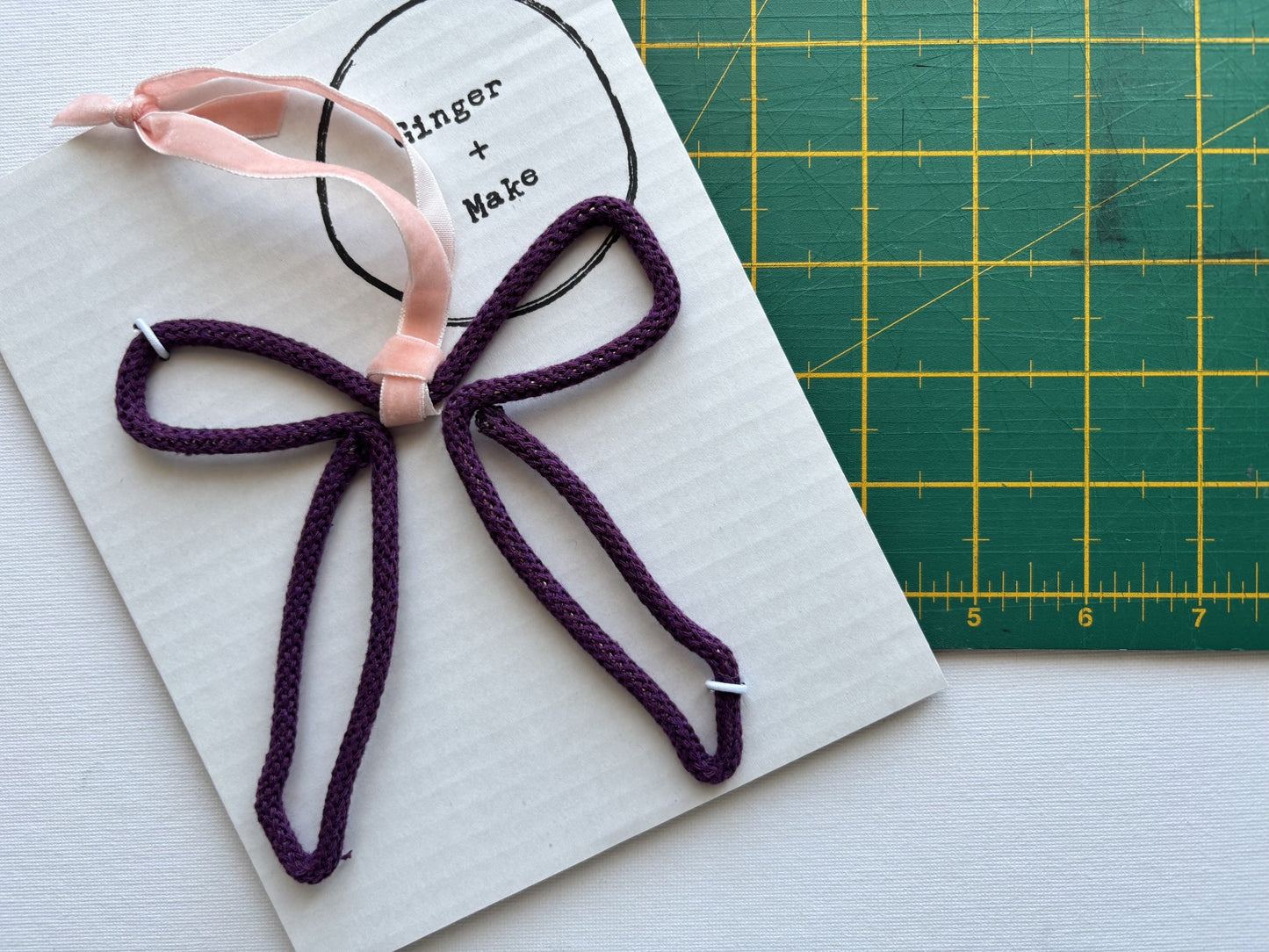 A handmade wire bow with dark purple braided cord, and a light pink ribbon loop, displayed on a white card next to a green cutting mat.