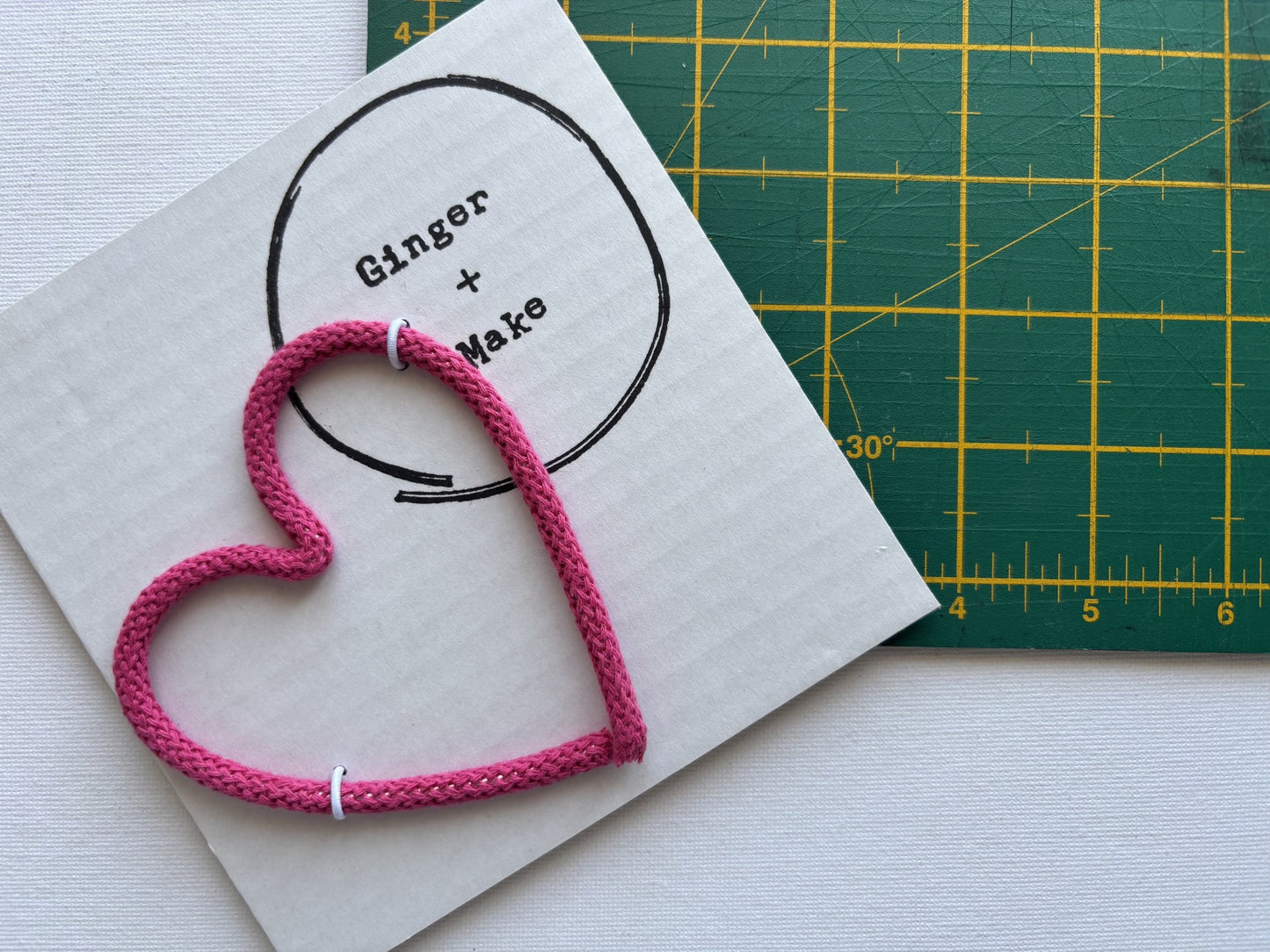A handmade wire heart with pink cord, displayed on white card printed with 'Ginger + Make' next to a green cutting mat.