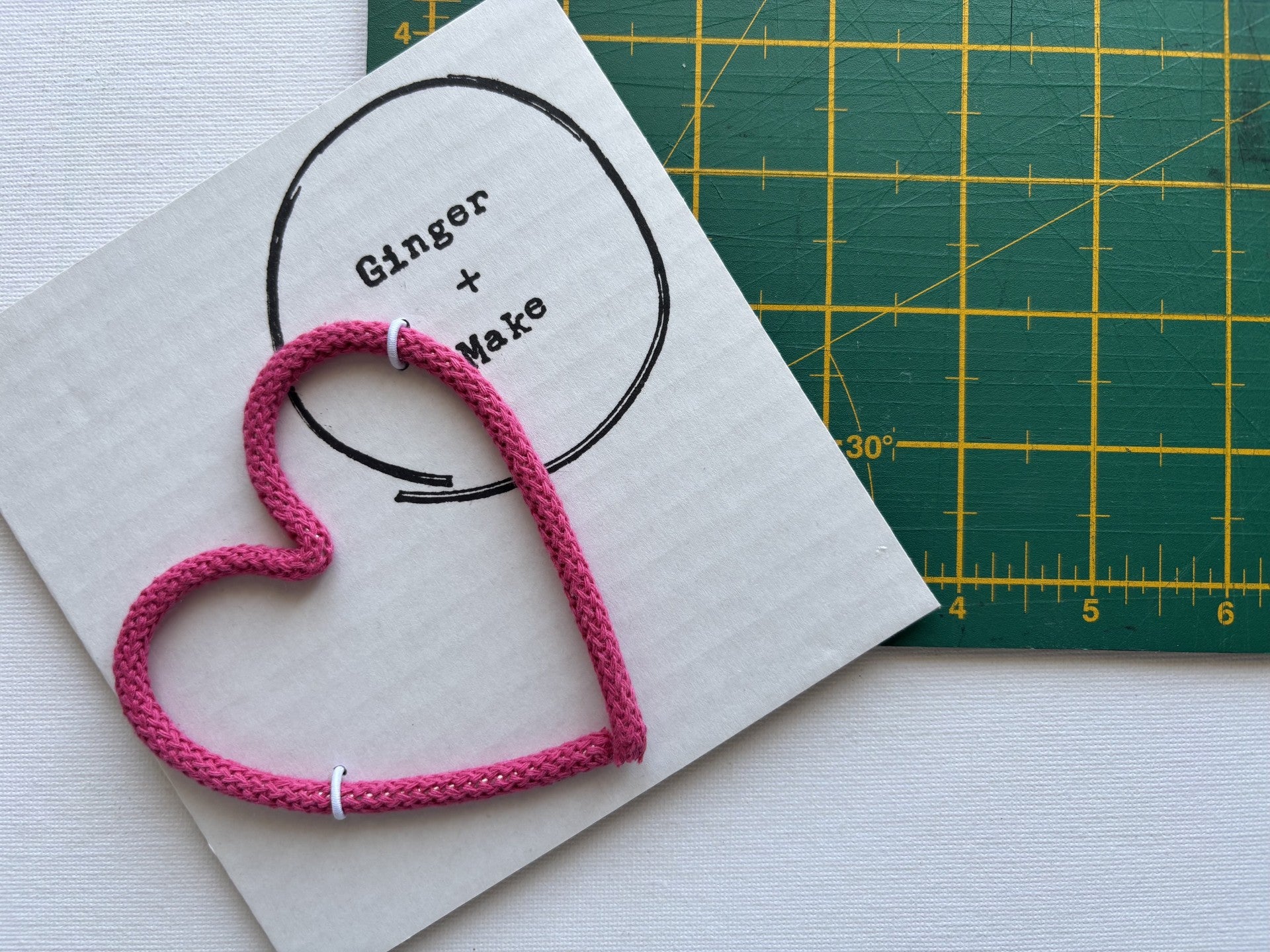 A handmade wire heart with pink cord, displayed on white card printed with 'Ginger + Make' next to a green cutting mat.