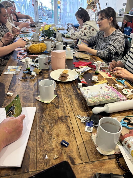 People sitting around a wooden table with various items including craft materials, cups and snacks. 