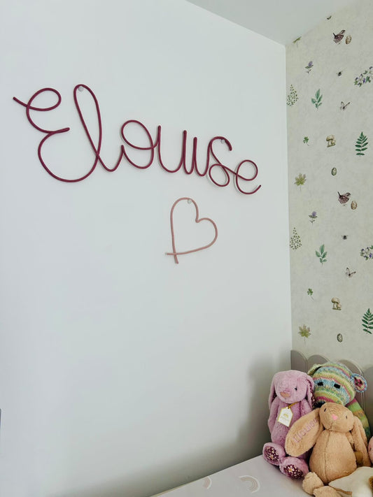 A custom wire wall sign spelling 'Elouse' in dark pink with a matching pink heart , mounted on a white wall in a child's bedroom next to patterned wallpaper and a pile of stuffed toys.