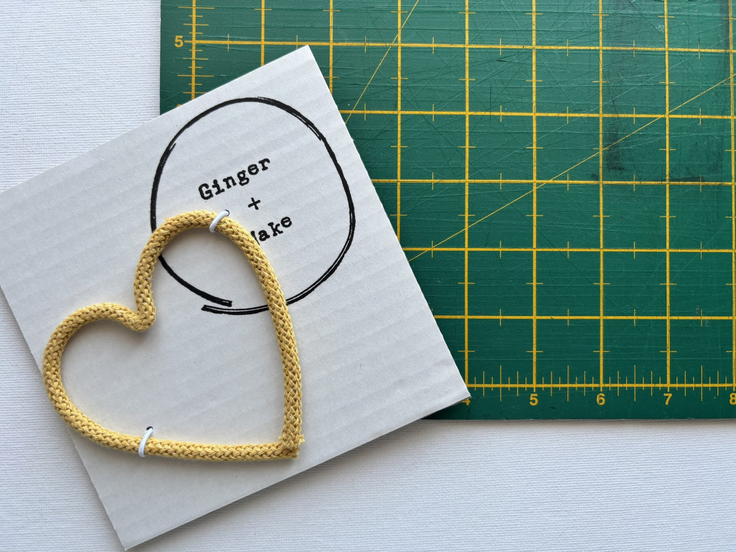 A handmade wire heart with yellow cord, displayed on white card printed with 'Ginger + Make' next to a green cutting mat.