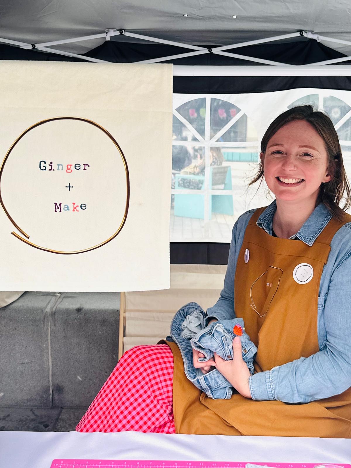 Jenni in a brown apron holding denim fabric under a tent with 'Ginger + Make' branding.