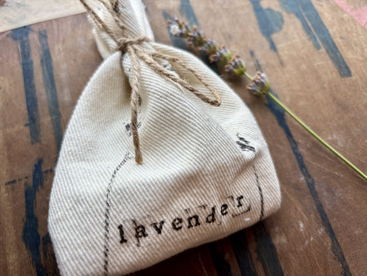 A small off-white fabric pouch printed with the word "lavender" in black script, tied with twine and placed on a rustic wooden surface next to a lavender sprig.