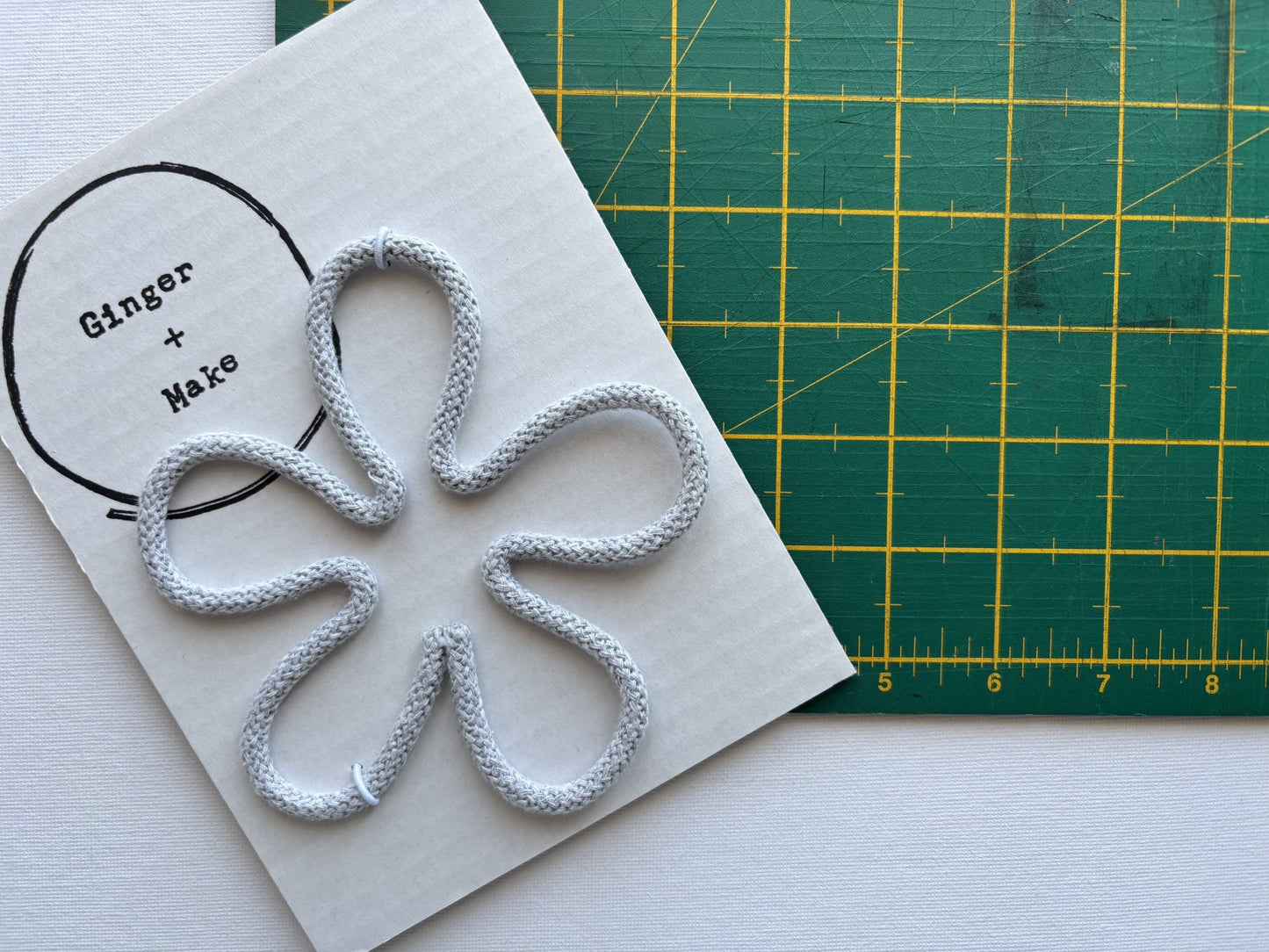 A handmade wire flower head with grey cord, displayed on white card printed with 'Ginger + Make' next to a green cutting mat.