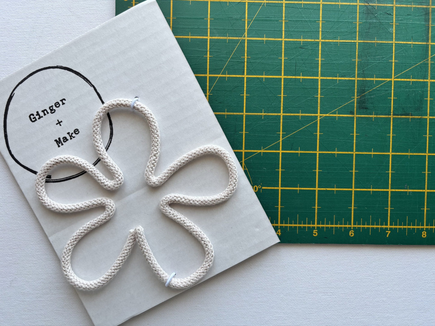 A handmade wire flower head with nude cord, displayed on white card printed with 'Ginger + Make' next to a green cutting mat.