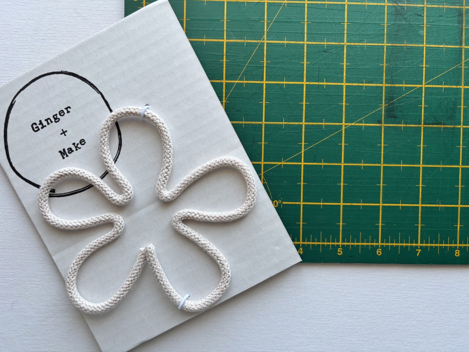 A handmade wire flower head with nude cord, displayed on white card printed with 'Ginger + Make' next to a green cutting mat.