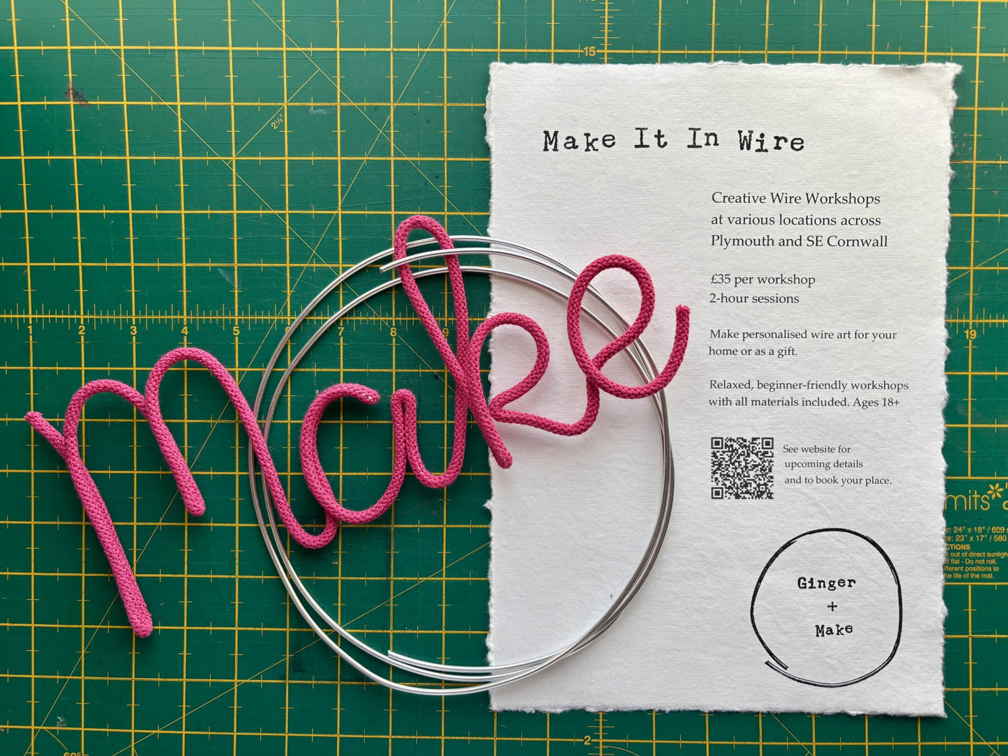 Pink wire spelling 'make' on a metal ring with a workshop flyer on a green cutting mat.