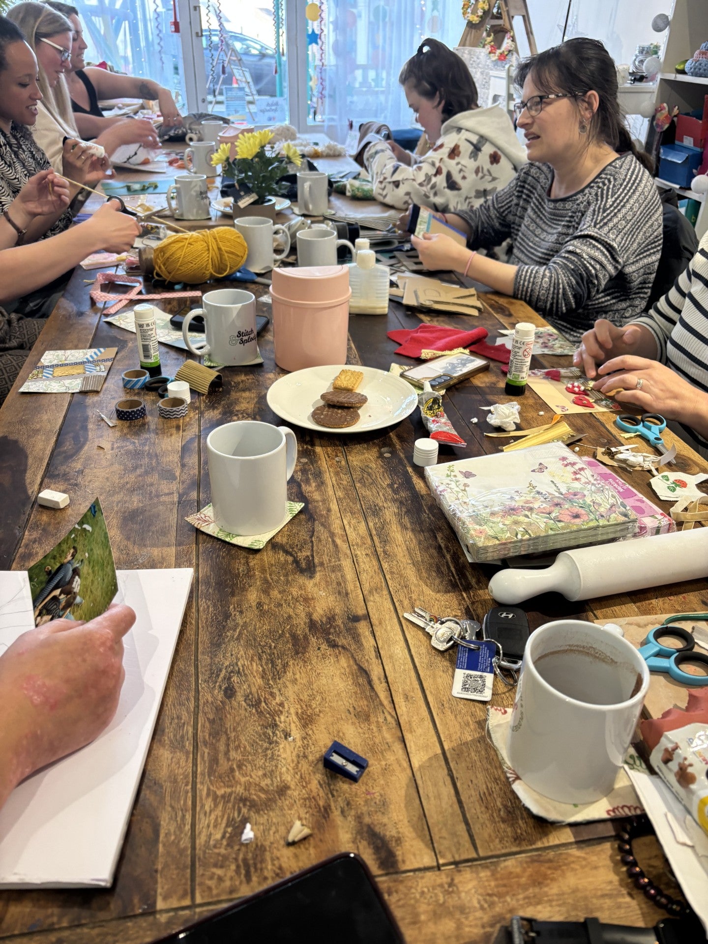 People sitting around a wooden craft table with various items including craft materials, cups and snacks.