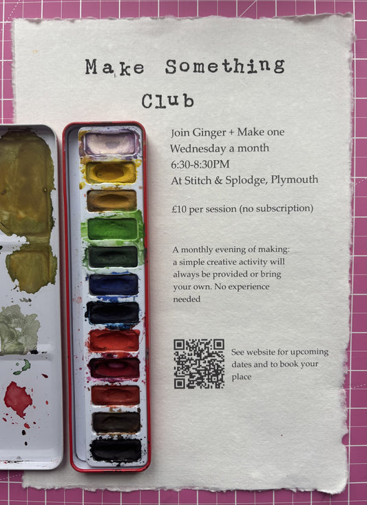 A printed invitation card for a 'Make Something Club' workshop, laid flat on a pink cutting mat background alongside an open watercolor paint palette with mixed colors.