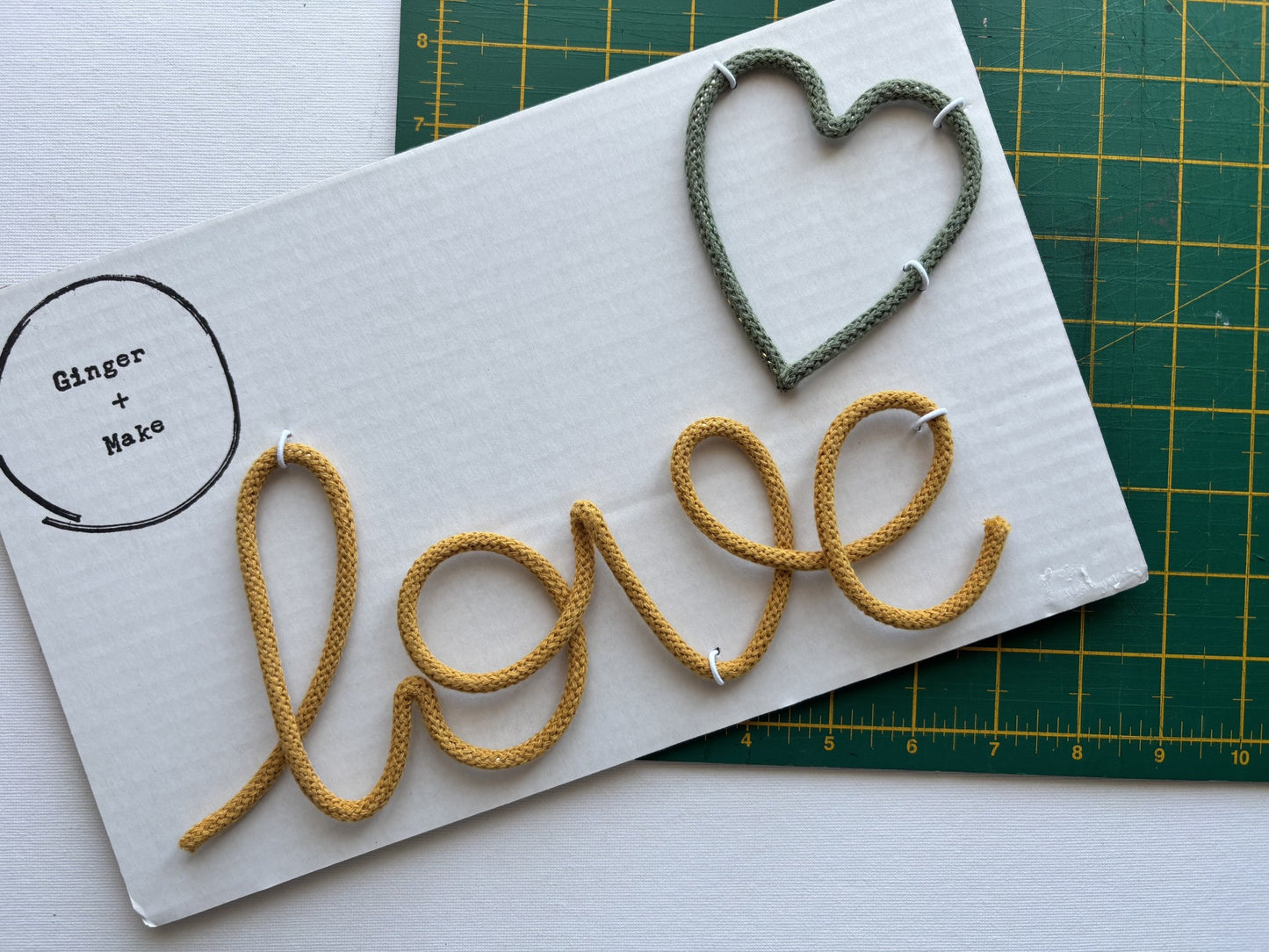 A white rectangular board with the word "love" spelled out in mustard cord and a heart outlined in green cord, placed on top of a green cutting mat. A circular logo reading "Ginger + Make" appears on the left side of the board.