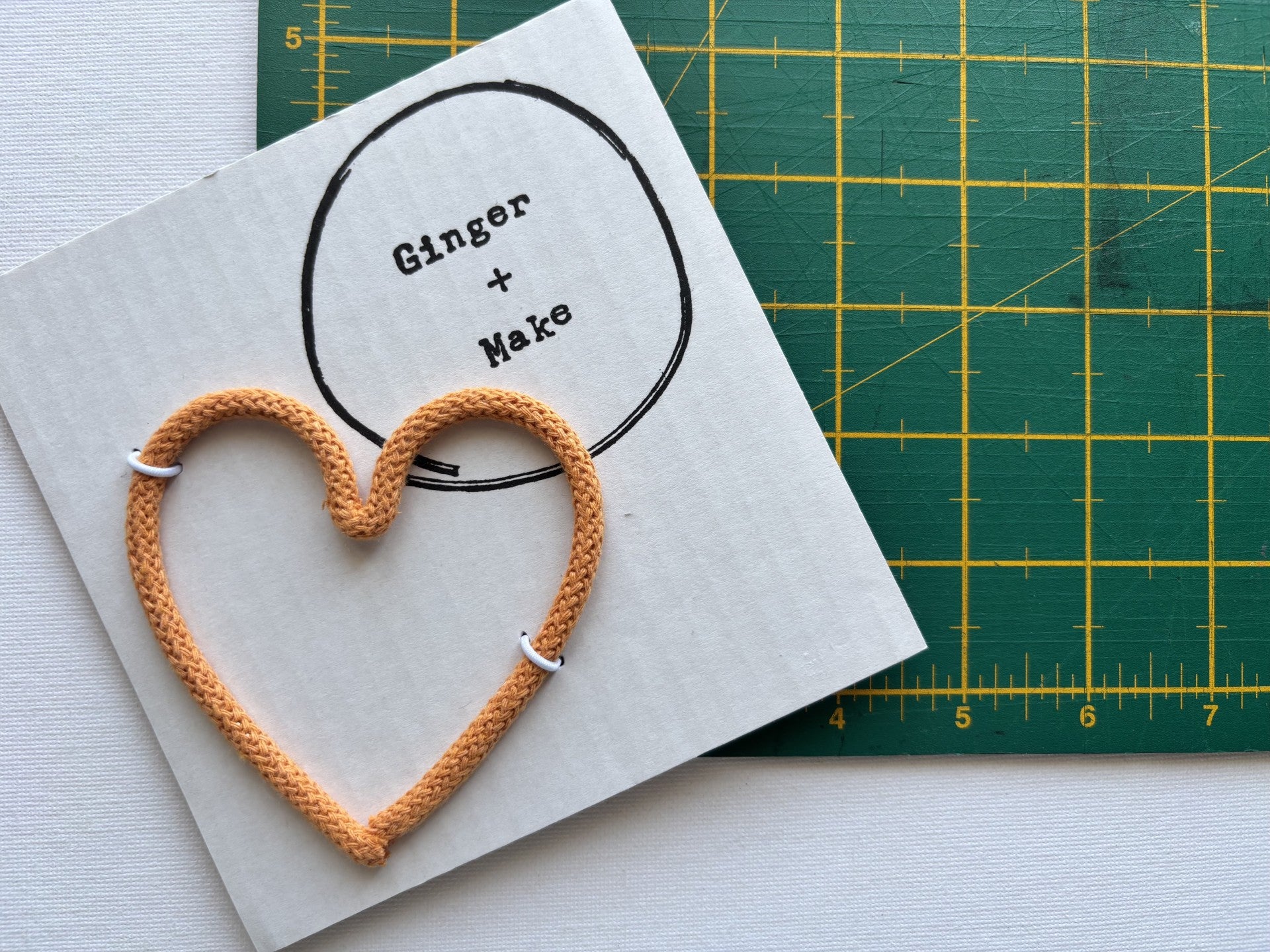 A handmade wire heart with orange cord, displayed on white card printed with 'Ginger + Make' next to a green cutting mat.