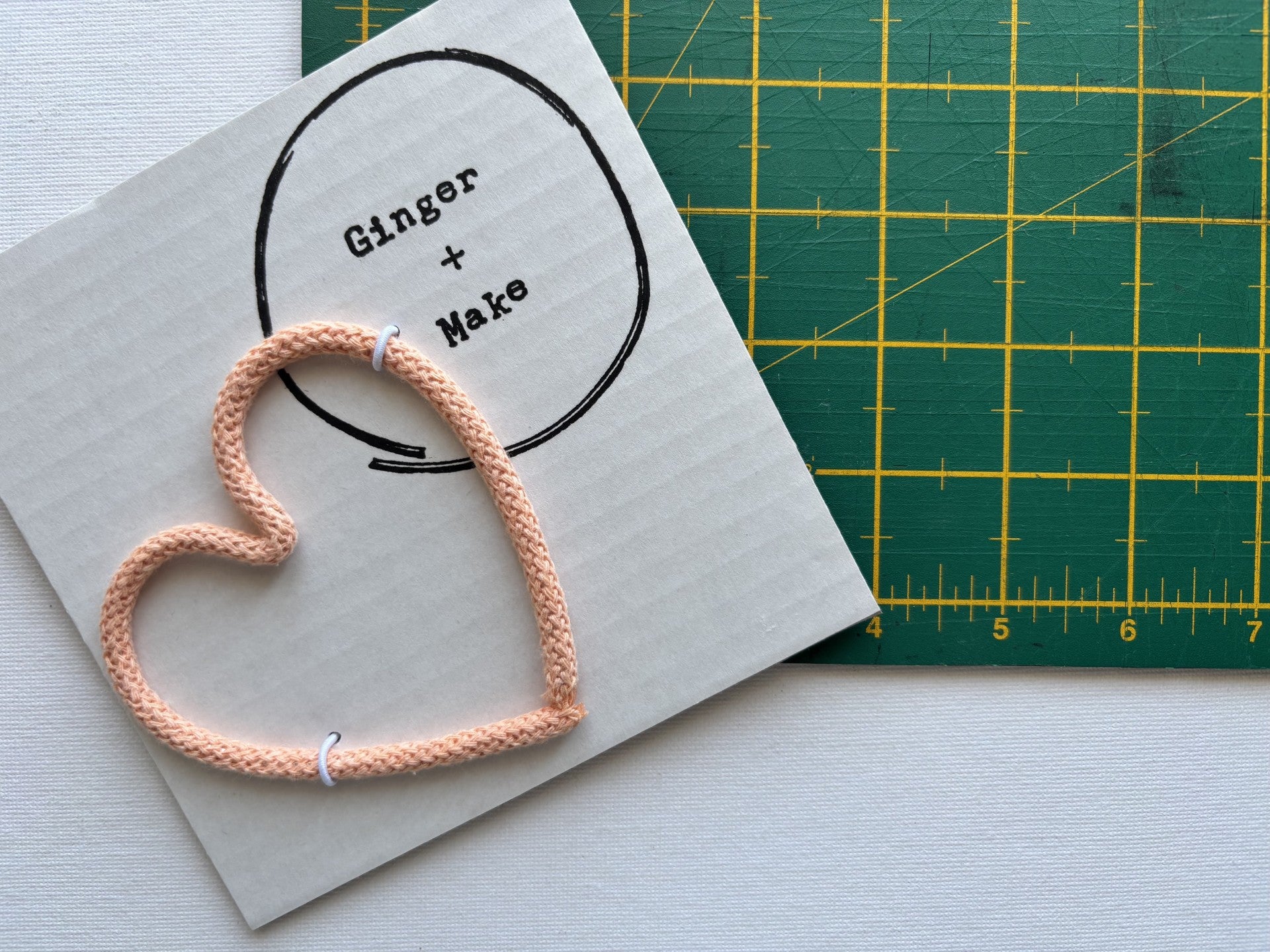 A handmade wire heart with peach cord, displayed on white card printed with 'Ginger + Make' next to a green cutting mat.