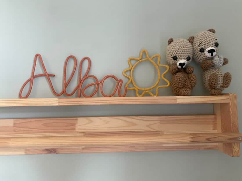 Wooden shelf with 'Alba' wire sign, sun-shaped wire figure, and two crocheted bears against a light gray wall.