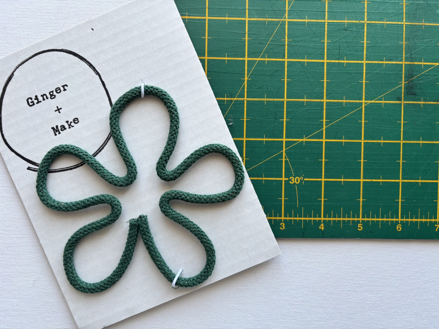 A handmade wire flower head with green cord, displayed on white card printed with 'Ginger + Make' next to a green cutting mat.