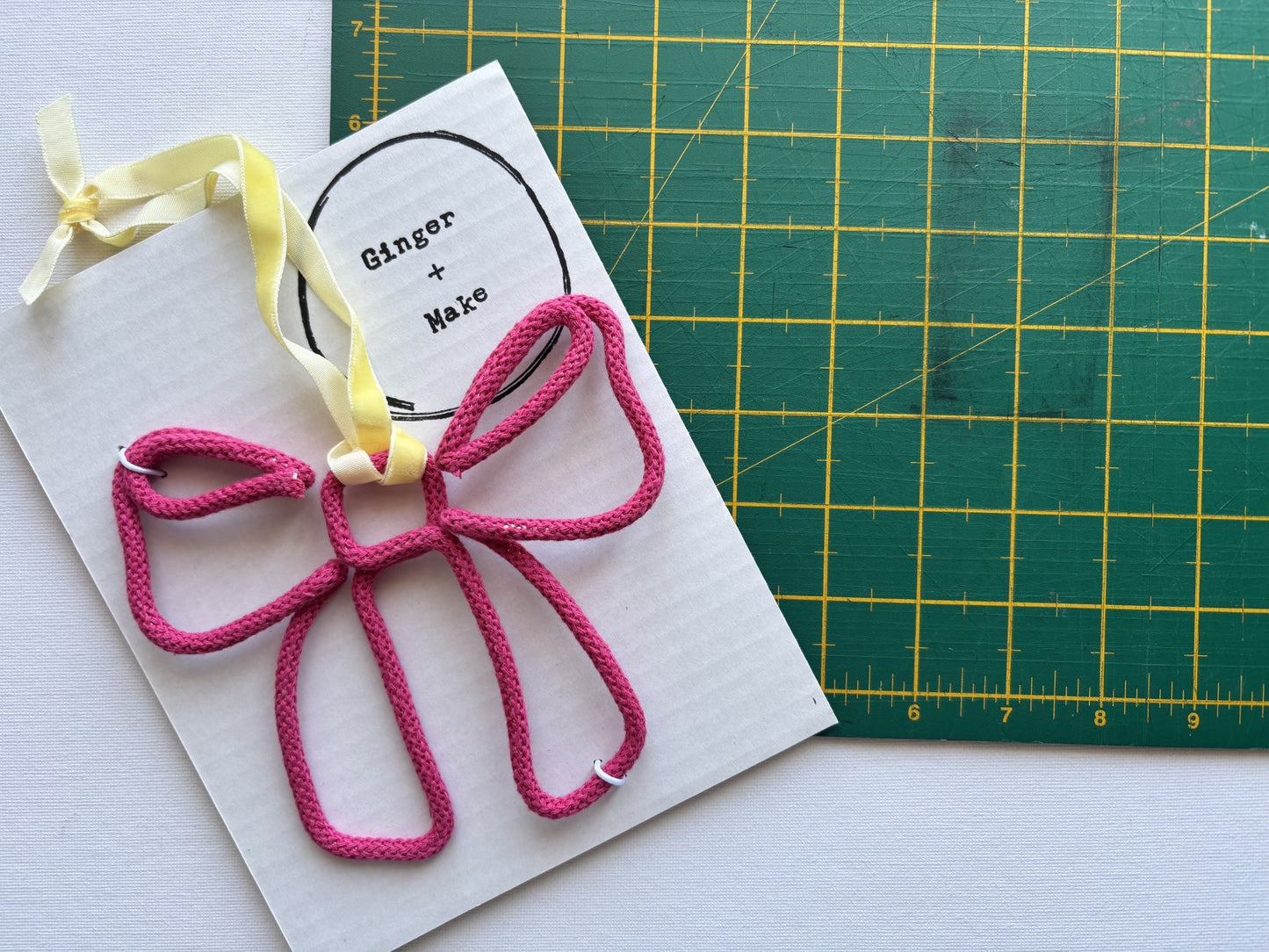A pink wire bow with a yellow ribbon loop, attached to a white card printed with 'Ginger + Make'. A green cutting mat is visible in the background.