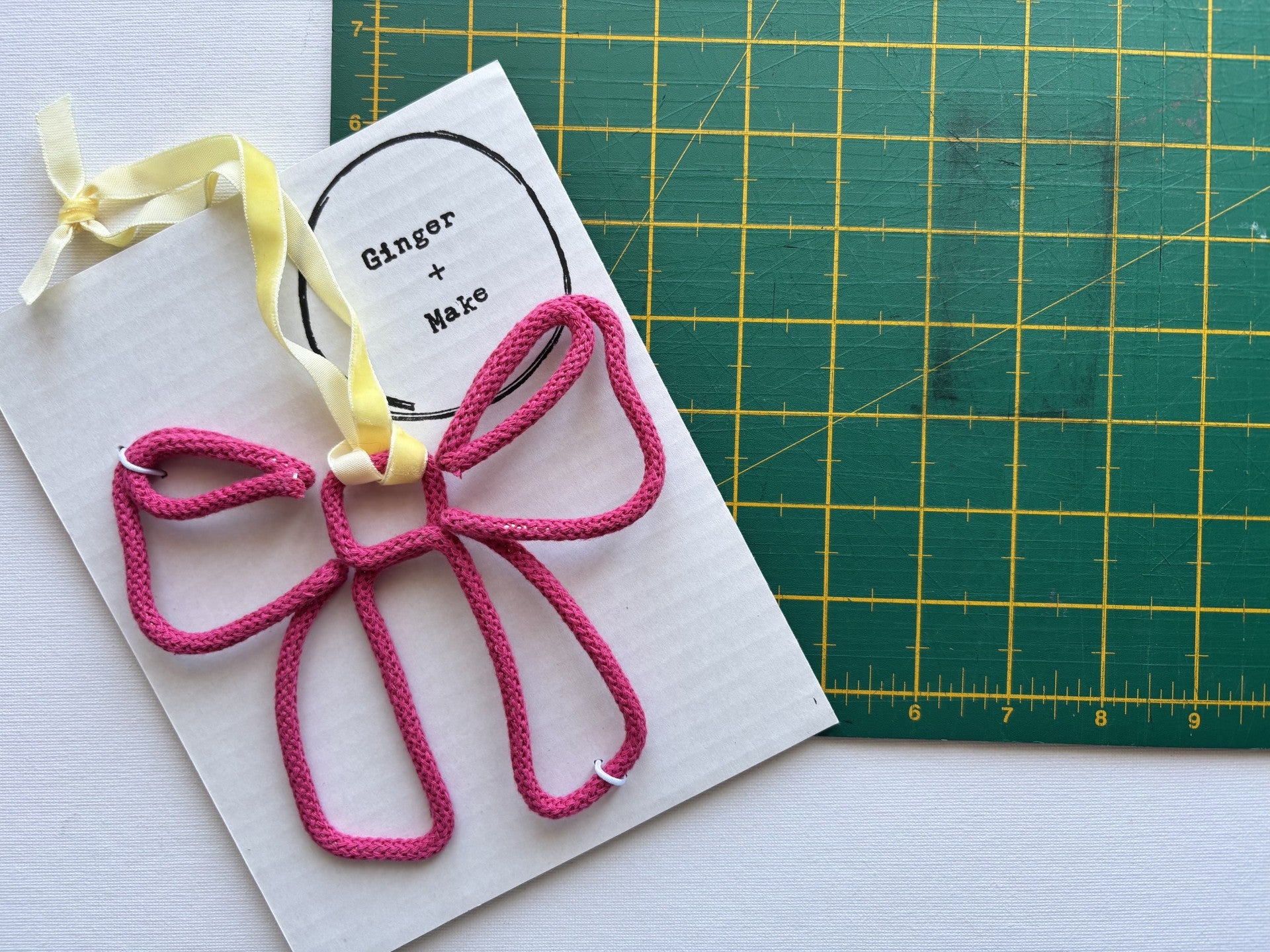 A pink wire bow with a yellow ribbon loop, attached to a white card printed with 'Ginger + Make'. A green cutting mat is visible in the background.