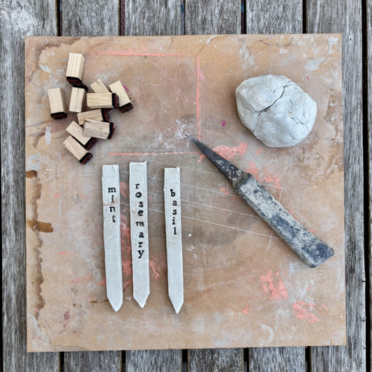 A top-down flat lay of gardening tools on a weathered wood surface: three white plant labels engraved with mint, rosemary, and basil, a small metal knife, several small wooden blocks, and a lump of pale clay on a tan work mat.