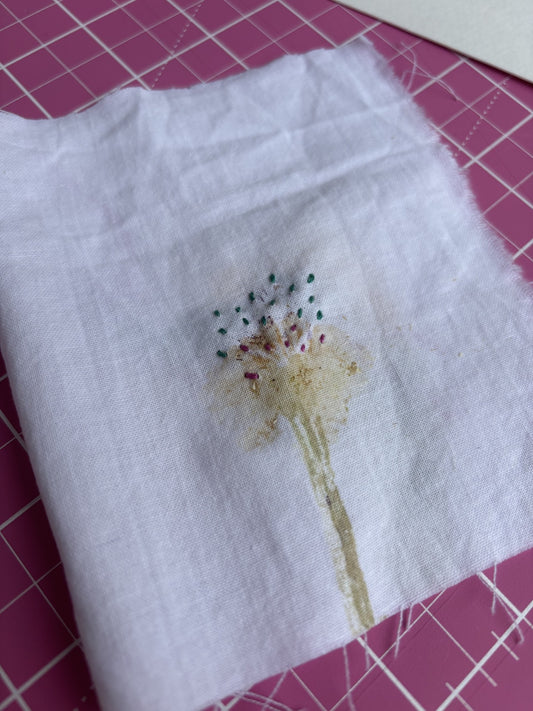 Embroidered dandelion design on a white fabric with pins marking stitches, on a pink grid background.