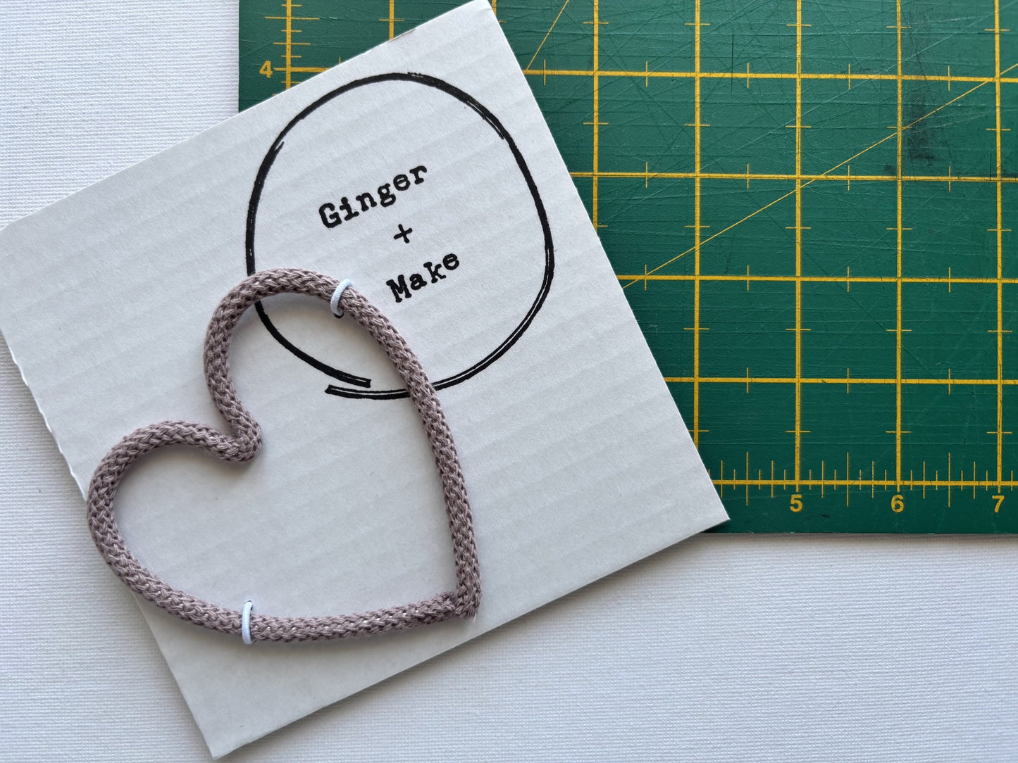 A handmade wire heart with mauve cord, displayed on white card printed with 'Ginger + Make' next to a green cutting mat.