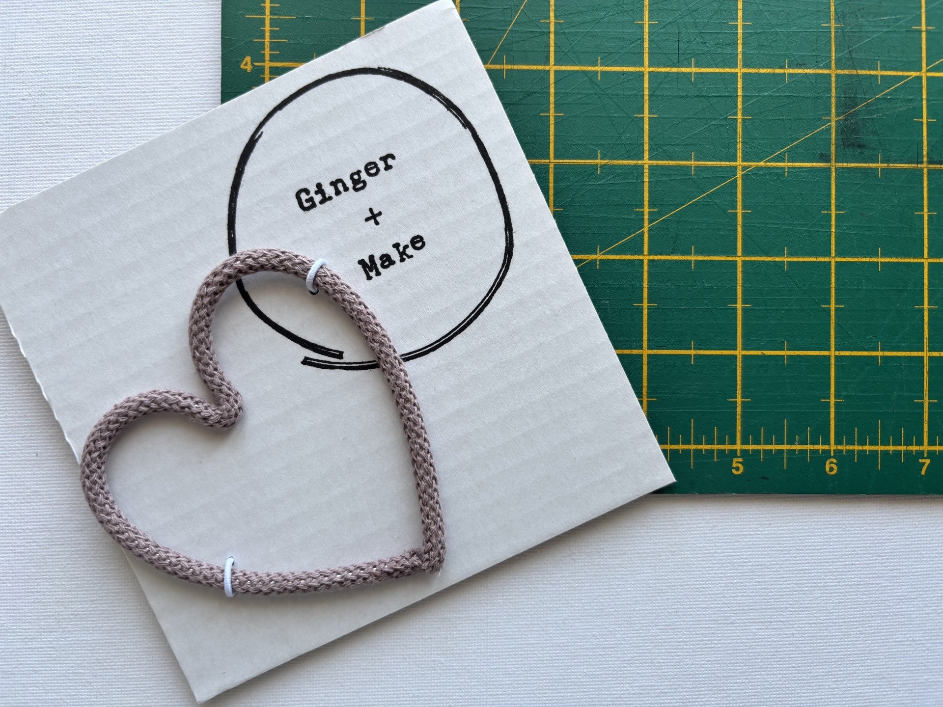 A handmade wire heart with mauve cord, displayed on white card printed with 'Ginger + Make' next to a green cutting mat.