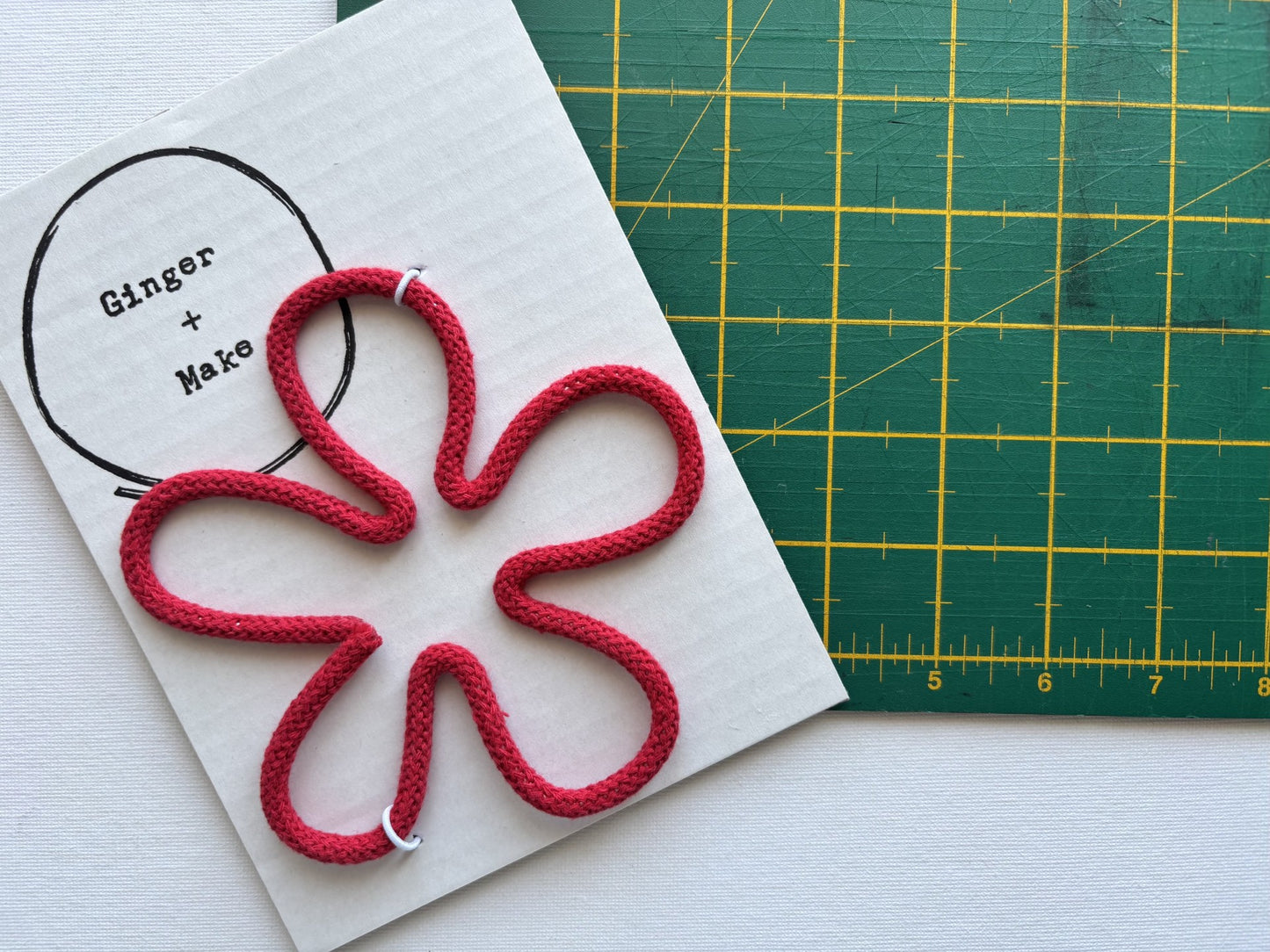A handmade wire flower head with red cord, displayed on white card printed with 'Ginger + Make' next to a green cutting mat.