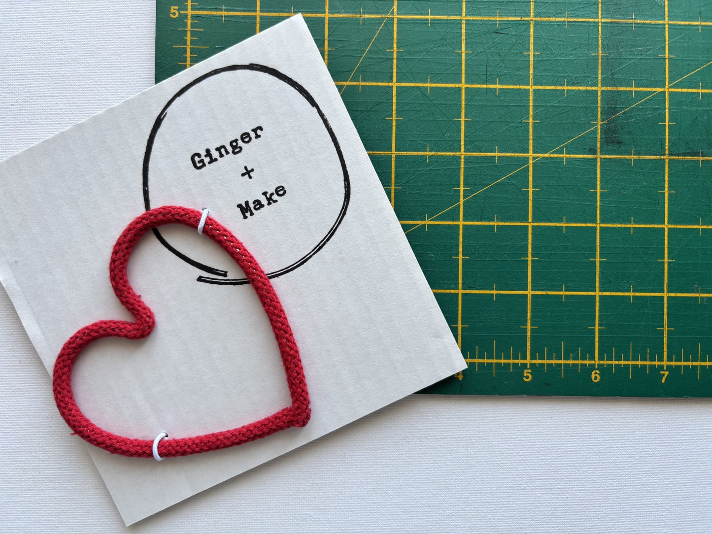 A red braided cord heart on white card printed with a circle and the words 'Ginger + Make', with a green cutting mat visible in the background.