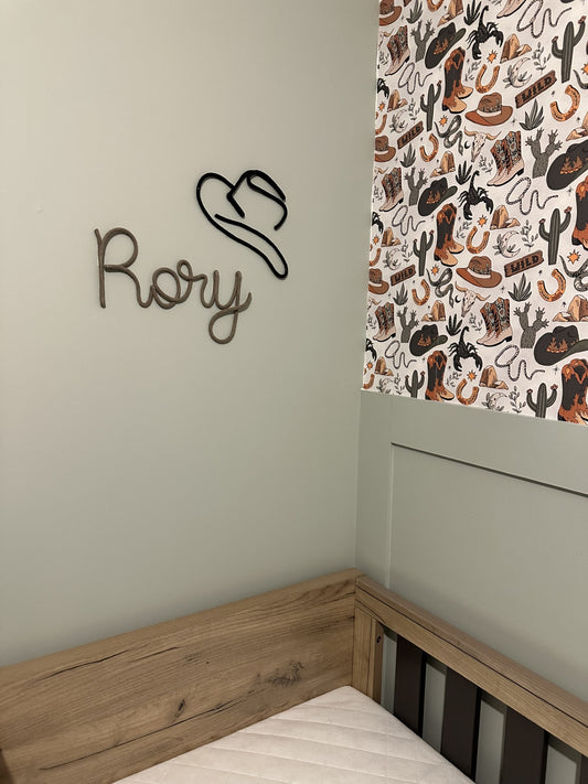A styled bedroom corner featuring a light wood bed frame with a white mattress, a sage green wall with a black wire cowboy hat and the name 'Rory' decoration, and a western-themed patterned wallpaper with boots, hats, and cacti.