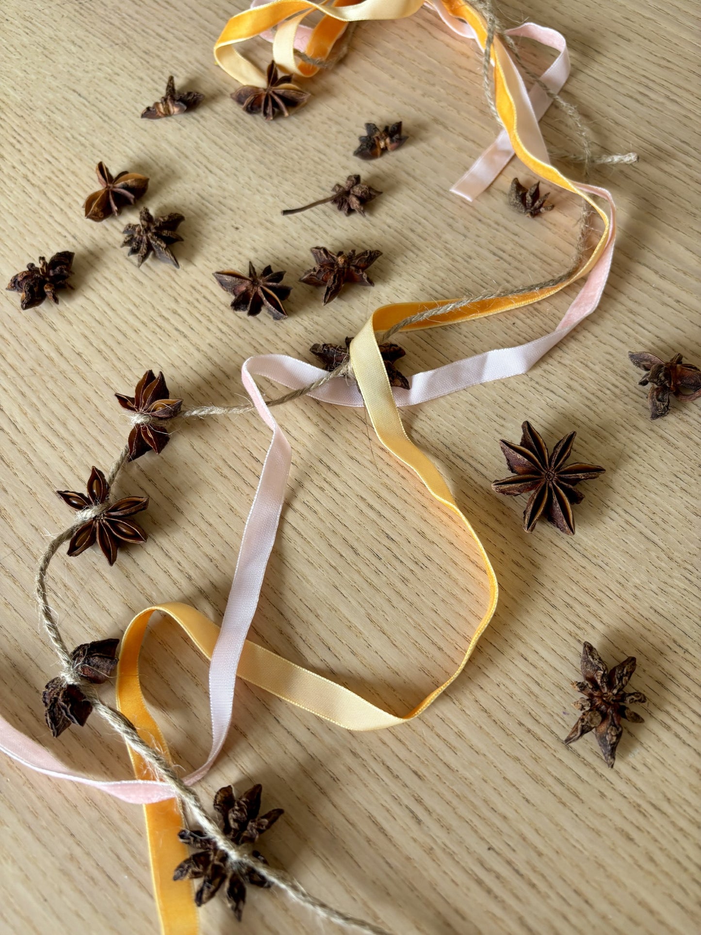 A garland of dried star anise spices threaded on twine with pastel yellow, pale pink, and cream ribbons, displayed on a light wood surface with additional star anise scattered around.