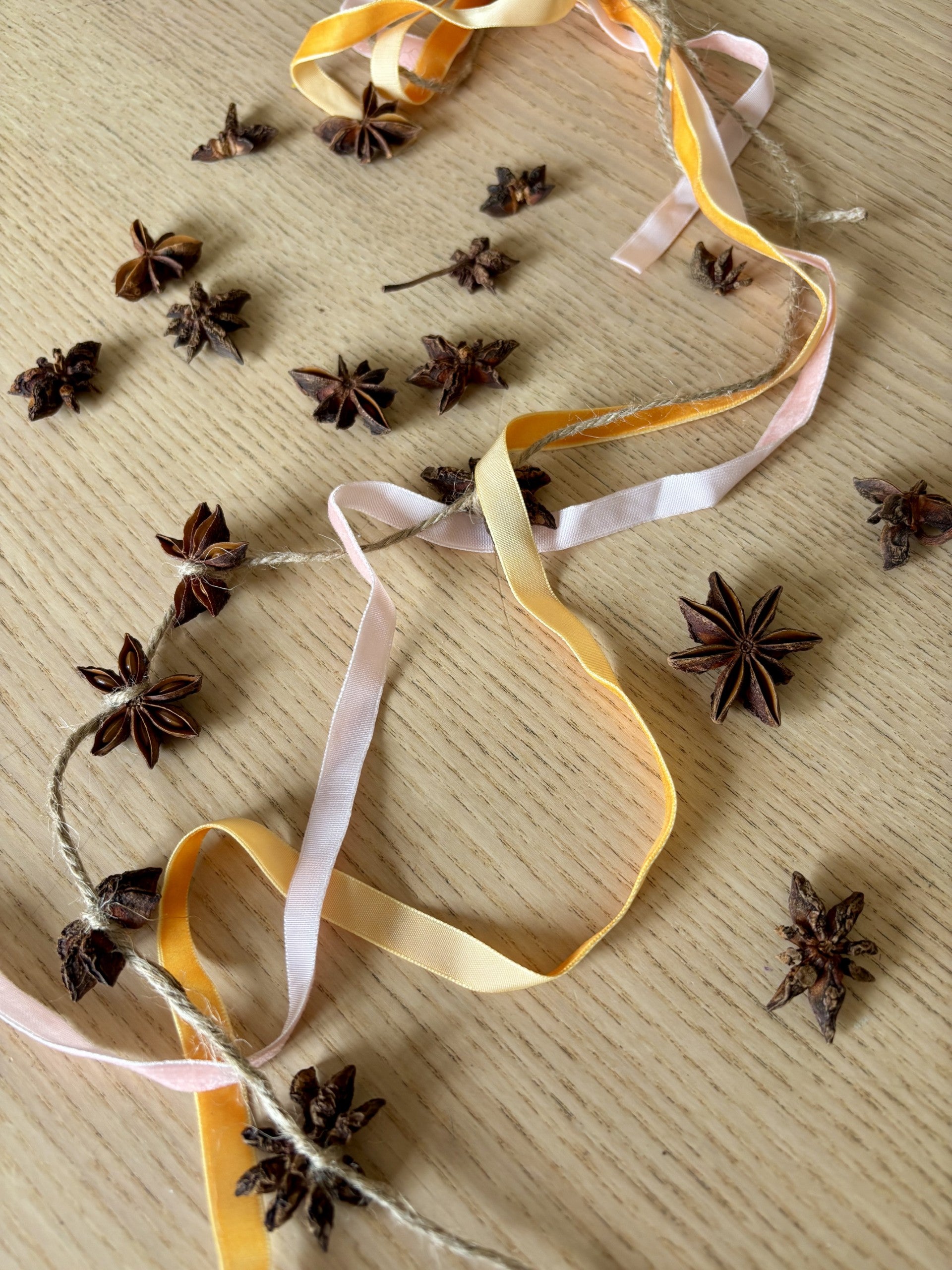 A garland of dried star anise spices threaded on twine with pastel yellow, pale pink, and cream ribbons, displayed on a light wood surface with additional star anise scattered around.
