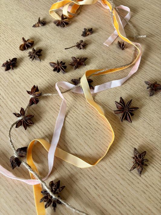 A garland of dried star anise spices threaded on twine with pastel yellow, pale pink, and cream ribbons, displayed on a light wood surface with additional star anise scattered around.