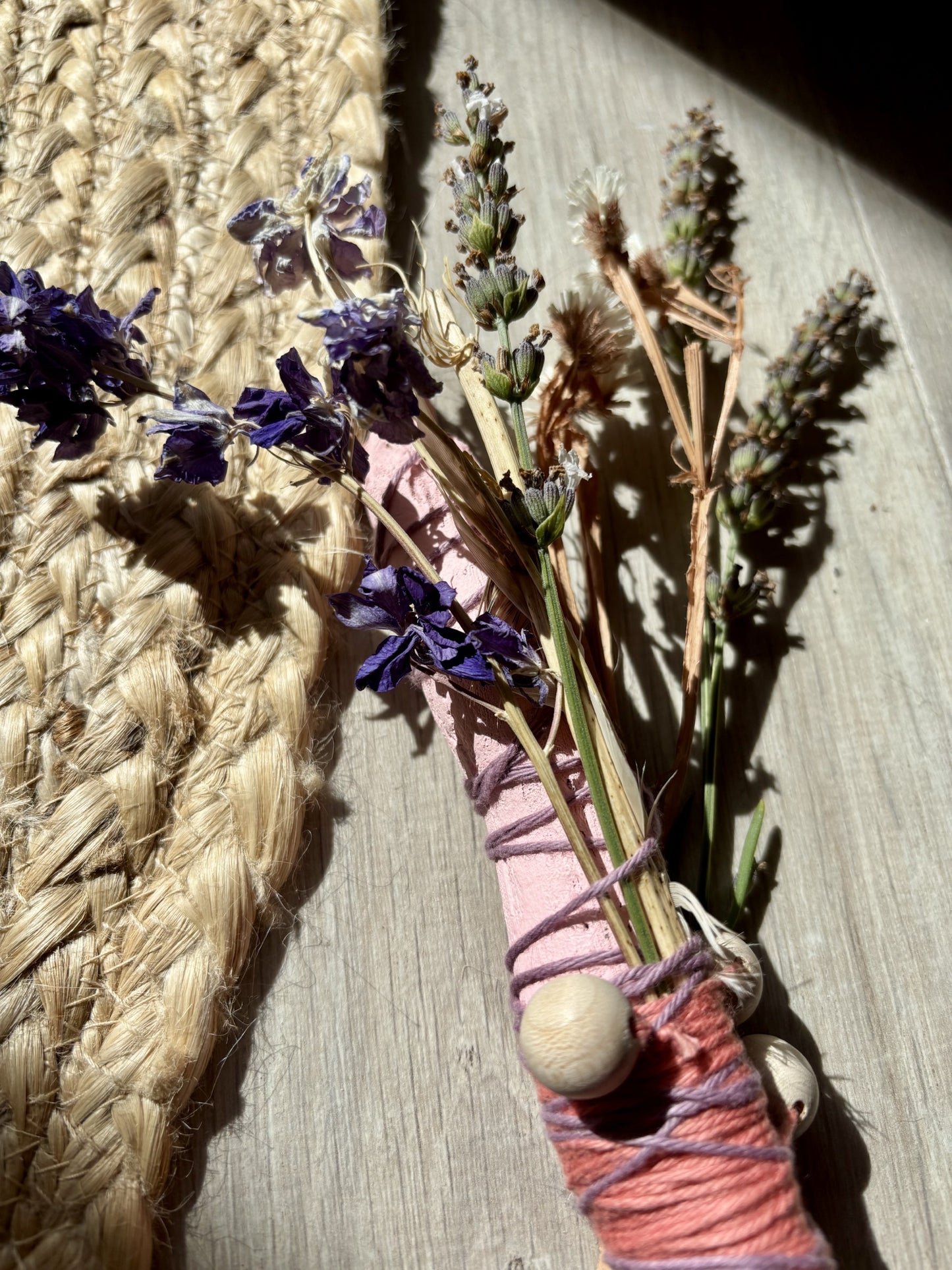 A close-up of a handmade wand wrapped with pink and purple yarn and adorned with wooden beads with a small bunch of dried lavender and grasses on a light wood surface.