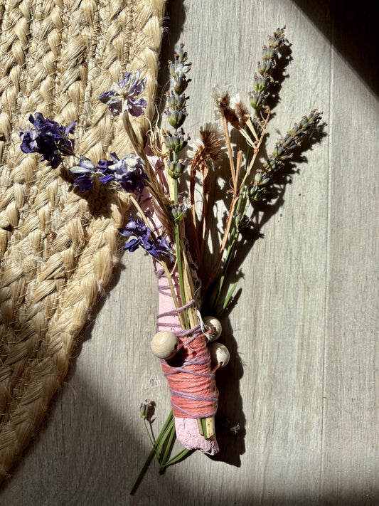 A dried flower and herb bundle with purple lavender, green stems, and pink wrapping, tied with twine and small wooden beads, placed on a light wood surface next to a woven straw mat.