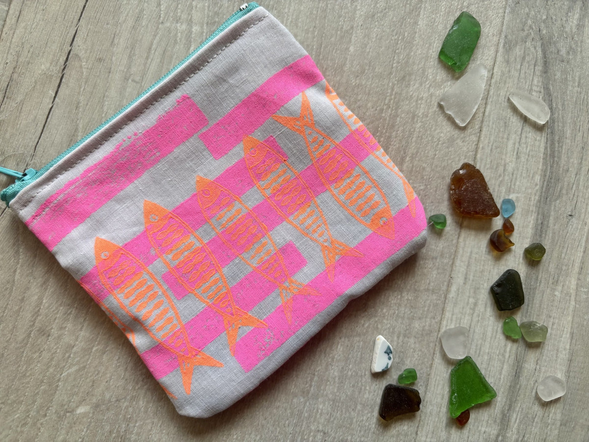 A fabric zippered pouch with a pink striped pattern and orange fish motifs, displayed on a light wood surface with scattered sea glass.