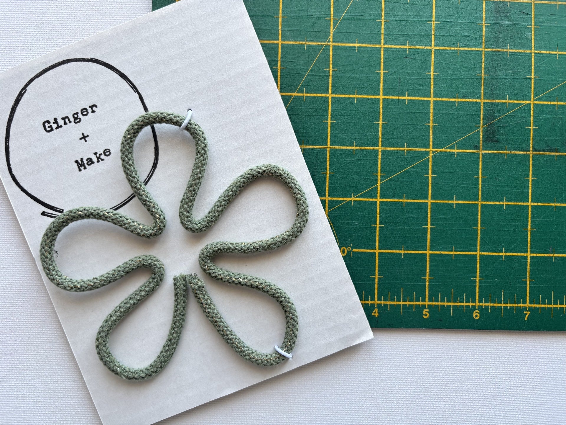 A handmade wire flower head with green cord, displayed on white card printed with 'Ginger + Make' next to a green cutting mat.