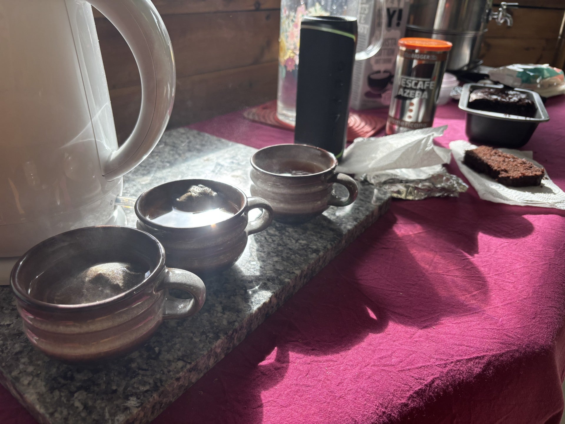 A tabletop scene with several small ceramic cups on a dark stone tray, alongside a white electric kettle, a black travel mug, a Nescafe coffee canister, and a piece of chocolate cake on a pink tablecloth.