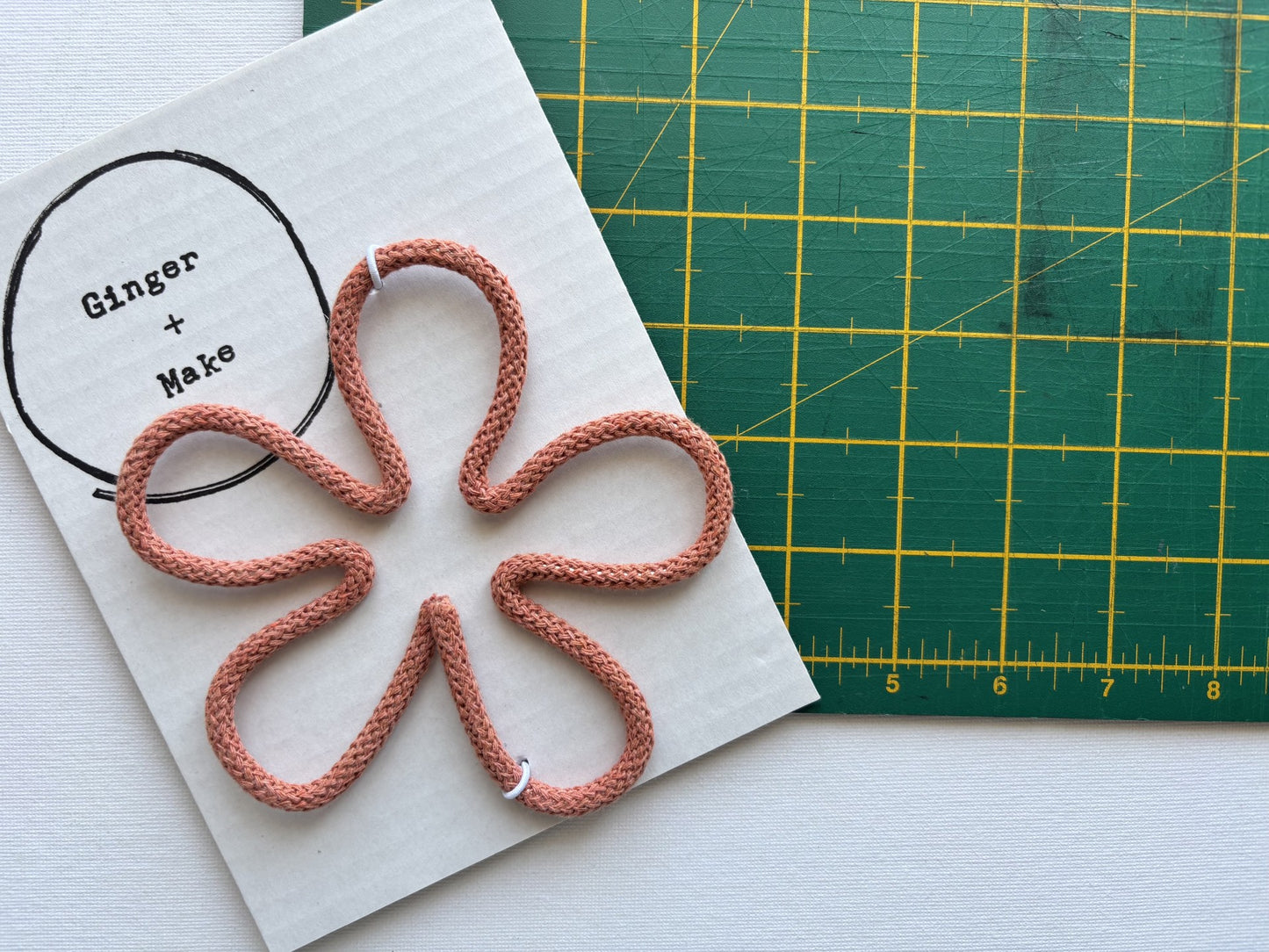 A handmade wire flower head with terracotta cord, displayed on white card printed with 'Ginger + Make' next to a green cutting mat.