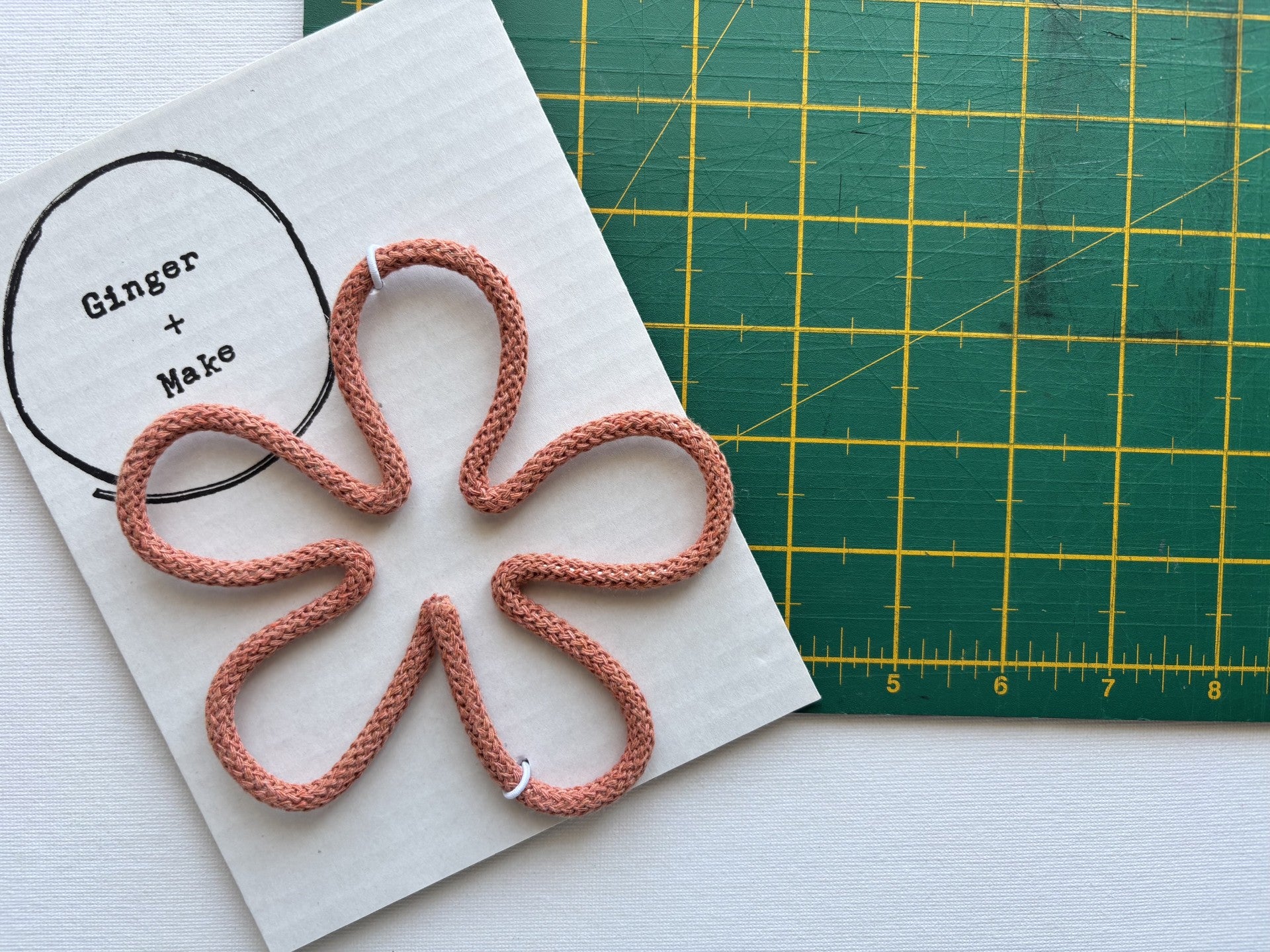 A handmade wire flower head with terracotta cord, displayed on white card printed with 'Ginger + Make' next to a green cutting mat.
