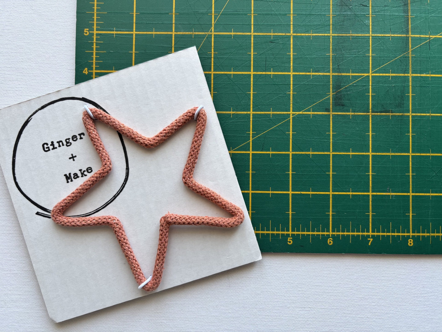 A terracotta braided cord shaped like a star, laid on top of white square card printed with the brand name 'Ginger + Make'. A green cutting mat with yellow grid lines is visible in the background.
