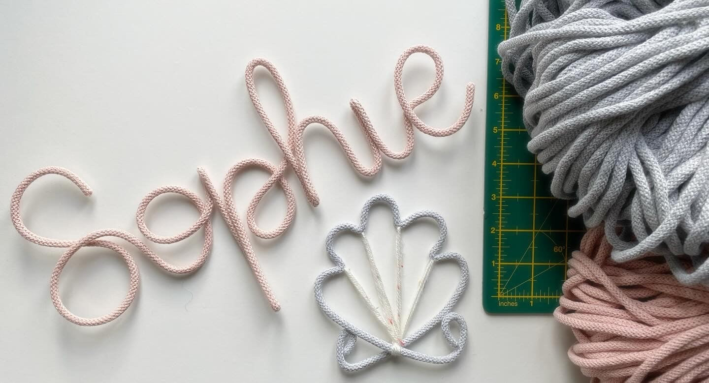A handmade wire name sign spelling out the name 'Sophie', made from aluminium wire and recycled cotton cord, displayed on a table alongside additional loose wires and a cutting mat.